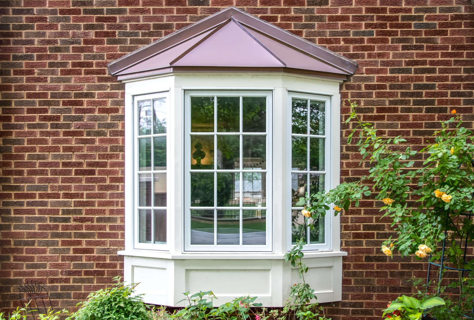 Bay window with copper roof on traditional brick home with flowers below and yellow roses extending out onto the window in springtime Bow & Bay Windows